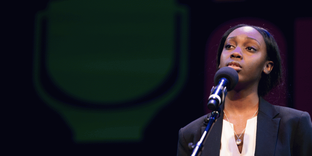 New York State Poetry Out Loud Champion 2019 Speaks into a mirophone on stage. They are wearing a blazer, white shirt, and stands against a dark backdrop.